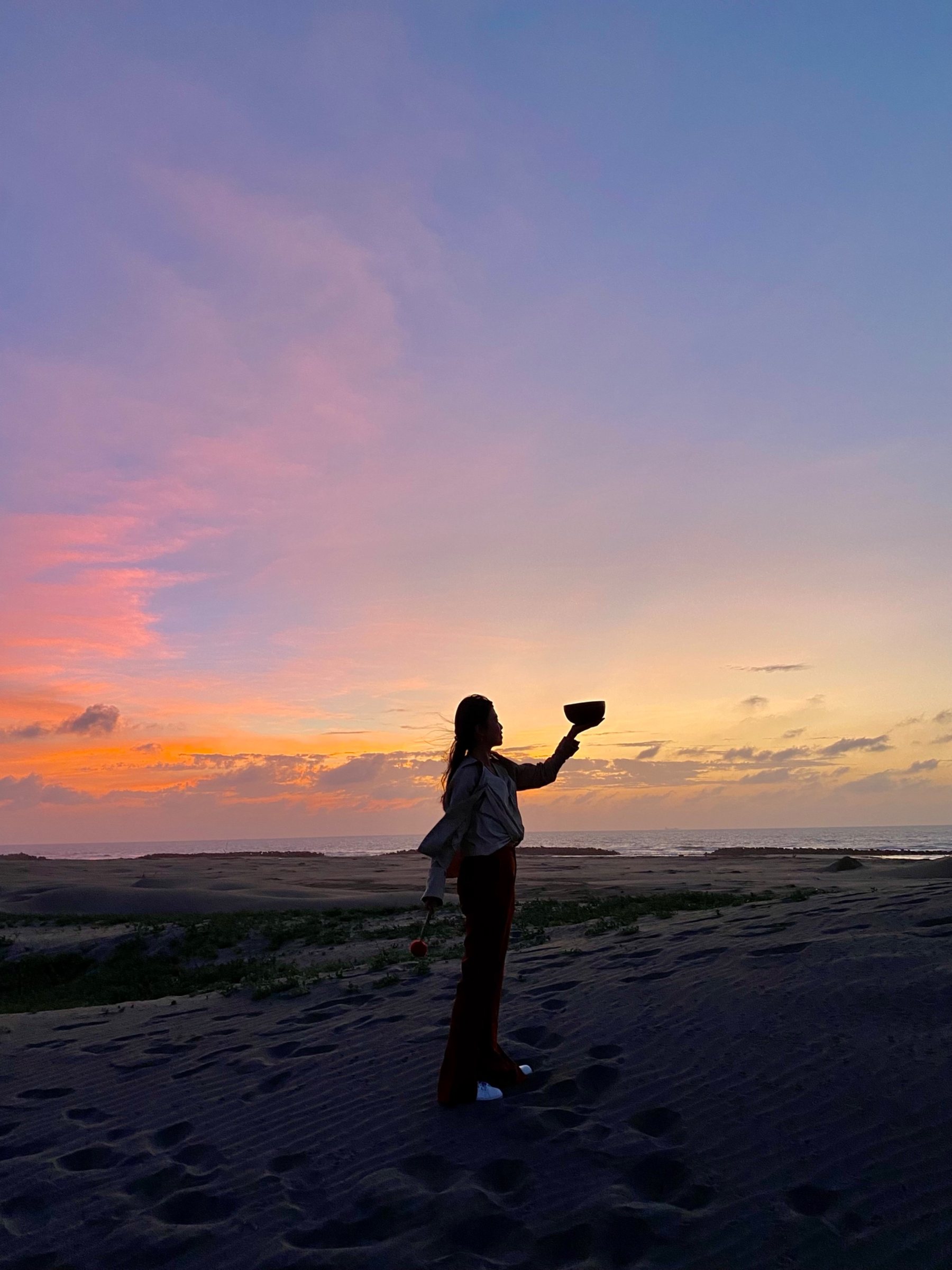 Helen raising a singing bowl to the sunset on the shore